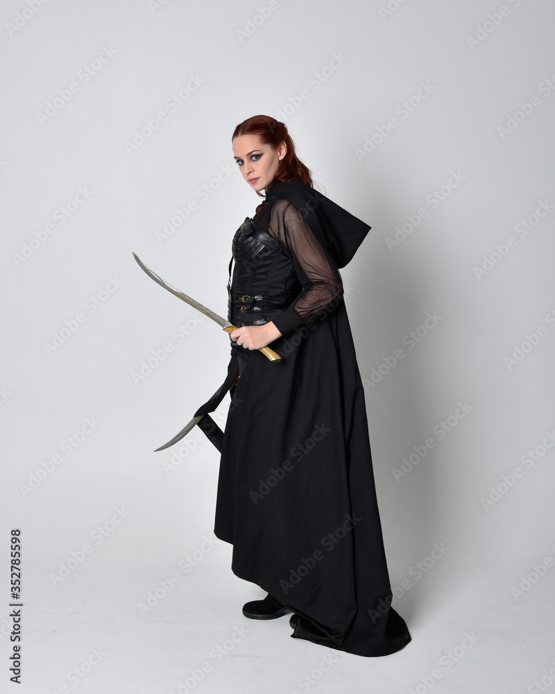 fantasy portrait of a woman with red hair wearing dark leather assassin costume with long black cloak. Full length standing pose holding a sword isolated against a studio background.