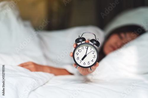 An asian woman holding and showing an alarm clock while sleeping on a white cozy bed in the morning