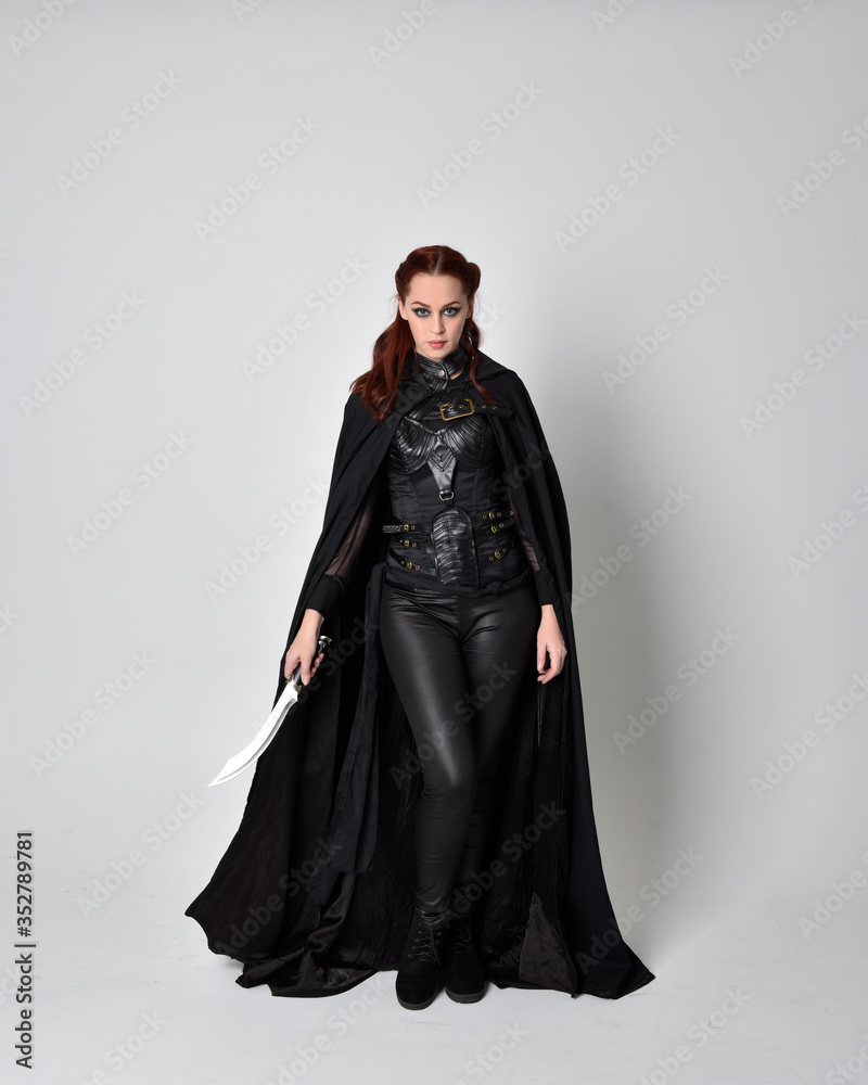 fantasy portrait of a woman with red hair wearing dark leather assassin  costume with long black cloak. Full length standing pose holding a sword  isolated against a studio background. Stock Photo