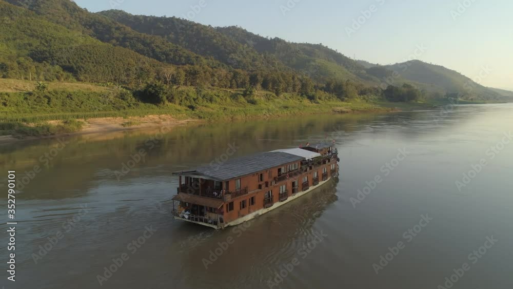Aerial panning shot of cruise ship with various flags moving on river against forest, drone panning near mountains against sky - Mekong River, Laos Aerial panning shot of cruise ship with various flags moving on river against forest, drone panning near mountains against sky - Mekong River, Laos