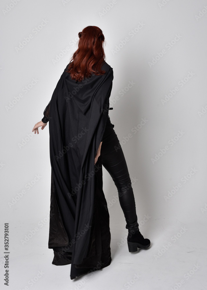 fantasy portrait of a woman wearing long black cloak. Full length standing pose  with back to the camera, isolated against a studio background.