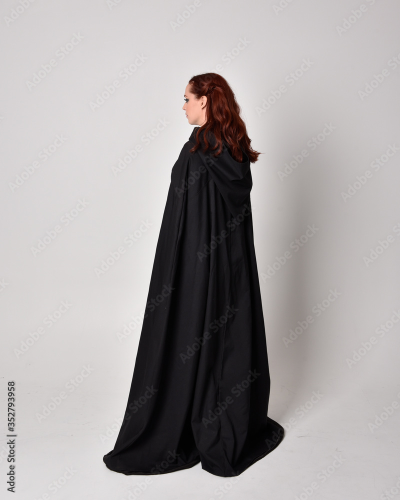 fantasy portrait of a woman wearing long black cloak. Full length standing pose  with back to the camera, isolated against a studio background.