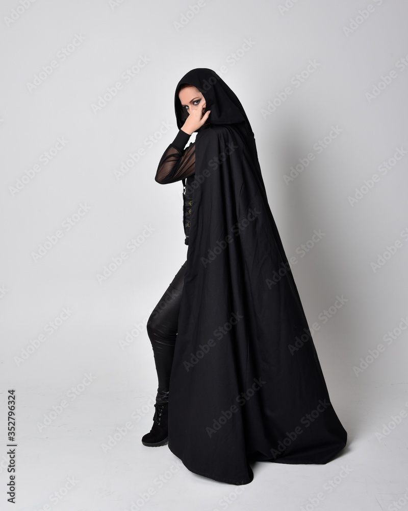 fantasy portrait of a woman with red hair wearing dark leather assassin costume with long black cloak. Full length standing pose in side profile isolated against a studio background. fantasy portrait of a woman with red hair wearing dark leather assassin costume with long black cloak. Full length standing pose in side profile isolated against a studio background.