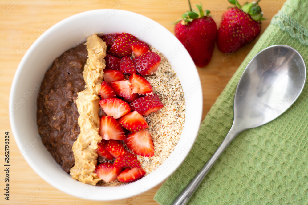 Chocolate cocoa oatmeal with almond meal, chopped strawberries and creamy peanut butter on a wooden surface. Strawberry, almonds, napkin and spoon in the background Chocolate cocoa oatmeal with almond meal, chopped strawberries and creamy peanut butter on a wooden surface. Strawberry, almonds, napkin and spoon in the background