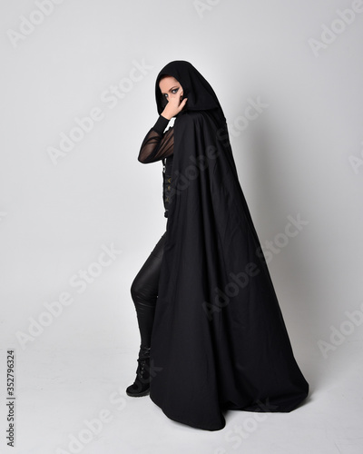 fantasy portrait of a woman with red hair wearing dark leather assassin costume with long black cloak. Full length standing pose in side profile isolated against a studio background.