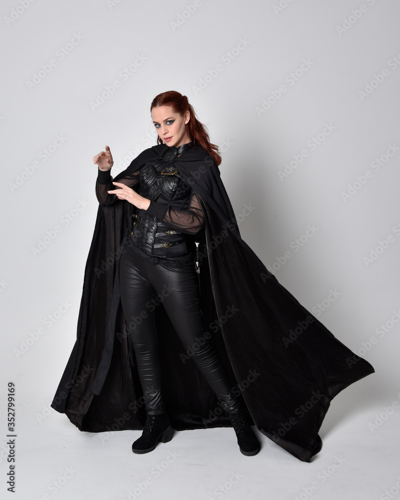 fantasy portrait of a woman with red hair wearing dark leather assassin costume with long black cloak. Full length standing pose  isolated against a studio background.