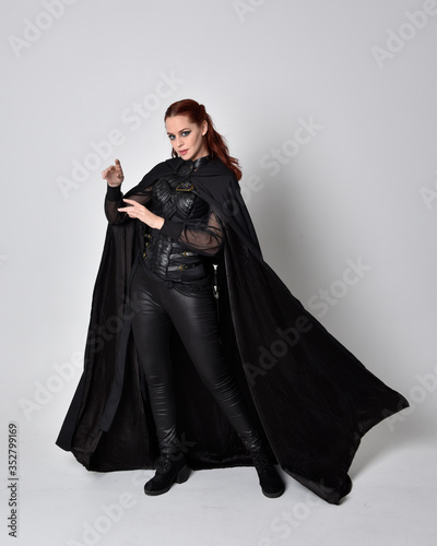fantasy portrait of a woman with red hair wearing dark leather assassin costume with long black cloak. Full length standing pose  isolated against a studio background.
