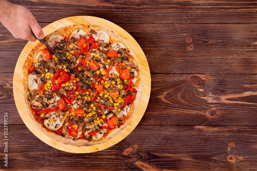 Man cut the pizza with a kitchen knife. Pizza with mushrooms, corn, cherry tomatos, courgettes and bell peppers on the wood platter which is on wooden rustic table, top view and copy space
