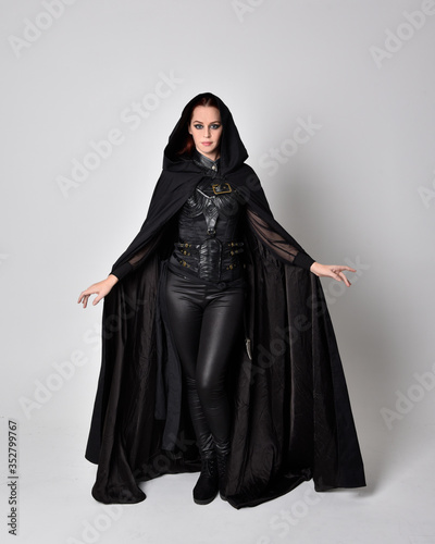 fantasy portrait of a woman with red hair wearing dark leather assassin costume with long black cloak. Full length standing pose  isolated against a studio background.
