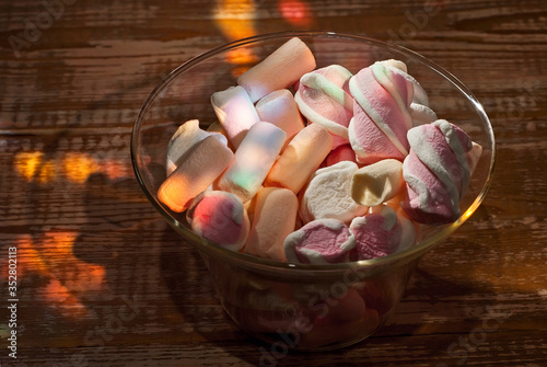 Sweets in a glass on a wooden table. Transparent utensils with marshmallows and biz on a shabby old board. Pink with white soft candies close up. photo