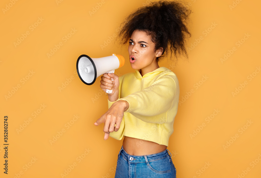 Girl shouts in megaphone and points forefinger to the side. Photo of african american girl on yellow background Girl shouts in megaphone and points forefinger to the side. Photo of african american girl on yellow background