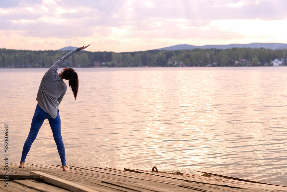 A charming athletic muscular woman works out at dawn on a wooden pier by a blue lake on a summer day