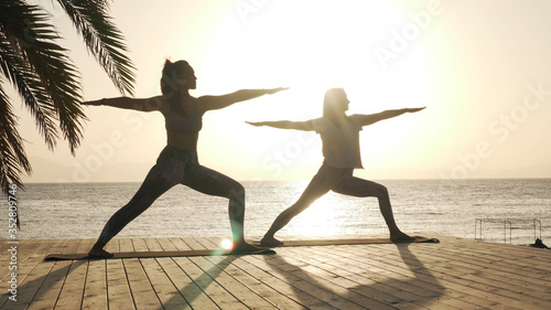 Silhouette of two women practice warrior yoga posture at seaside in sunshine
