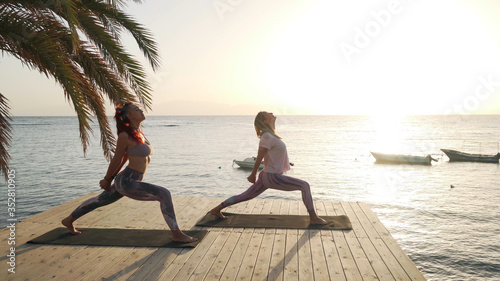 Two women practice warrior yoga posture in seaside pier at sunset