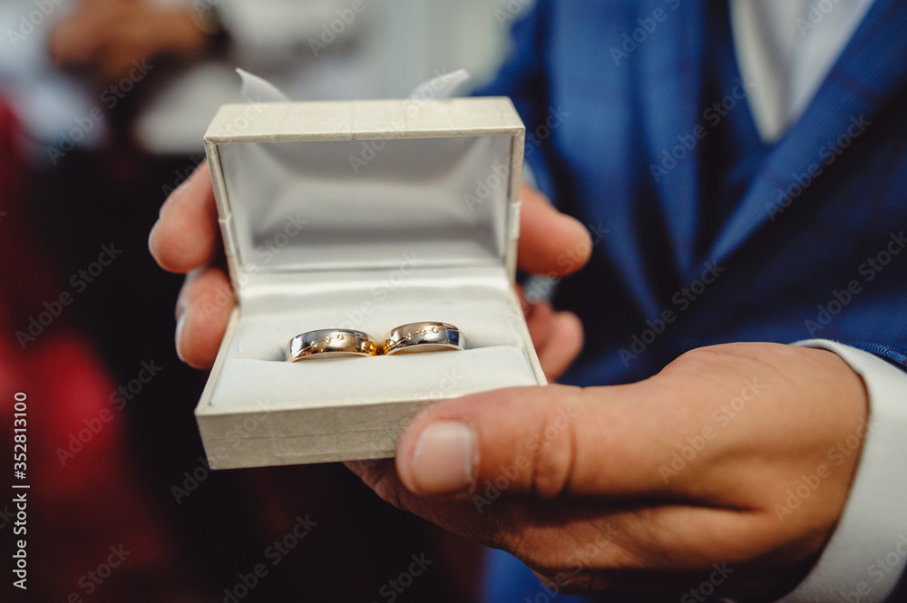 Fototapeta premium The bridegroom in a blue tuxedo holds in his hand the wedding rings. Close- up. Morning of the groom. Wedding accessories. Men's hands holding a box with rings. Film noise