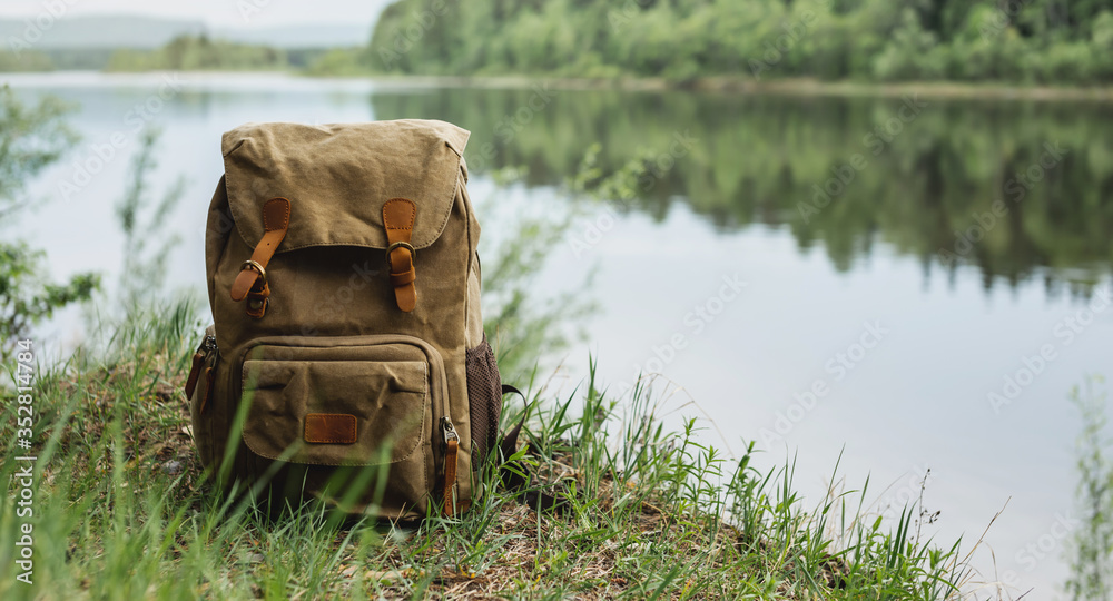 Obraz premium Tourist's brown backpack on the shore of a river. A great trip to nature in a dense green forest with a reservoir