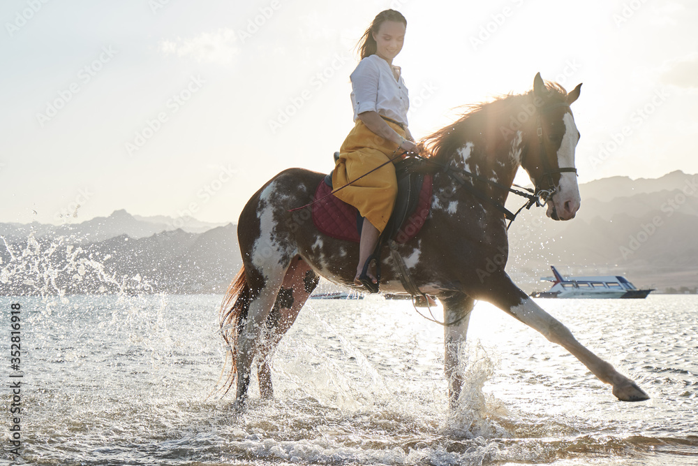 Caucasian blonde girl riding a horse through the water at sunrise Caucasian blonde girl riding a horse through the water at sunrise