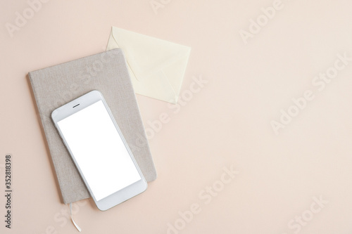 Flat lay, top view office desk table with mobile phone mockup with blank white screen, paper notebook and envelope on beige background. Business and technology concept.