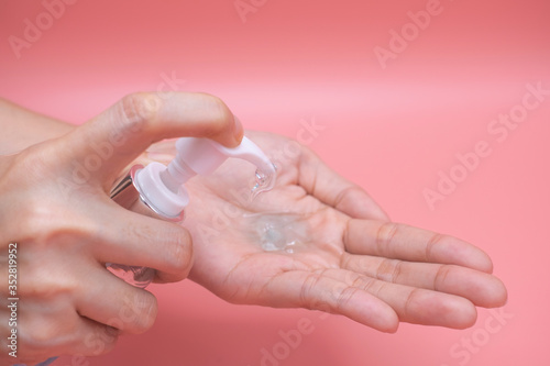 Woman hands using wash hand sanitizer gel pump dispenser. Clear sanitizer in pump bottle, for killing germs, bacteria and virus, pink background.