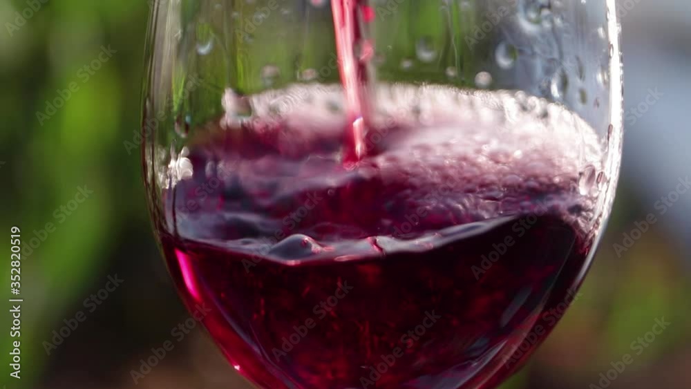 Close-up of pouring red wine into glass with green background. Real time