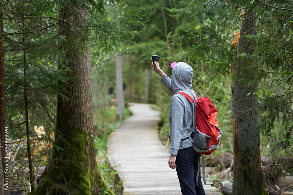 Travel. Young girl traveler with orange backpack walking on a wooden bridge in nature reserve and shooting video with an action camera.