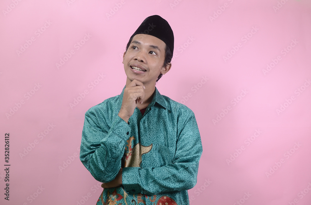 Fototapeta premium The young man seemed to think and look up. Portrait of young Asian man wearing batik shirt and skullcaps isolated on a pink background