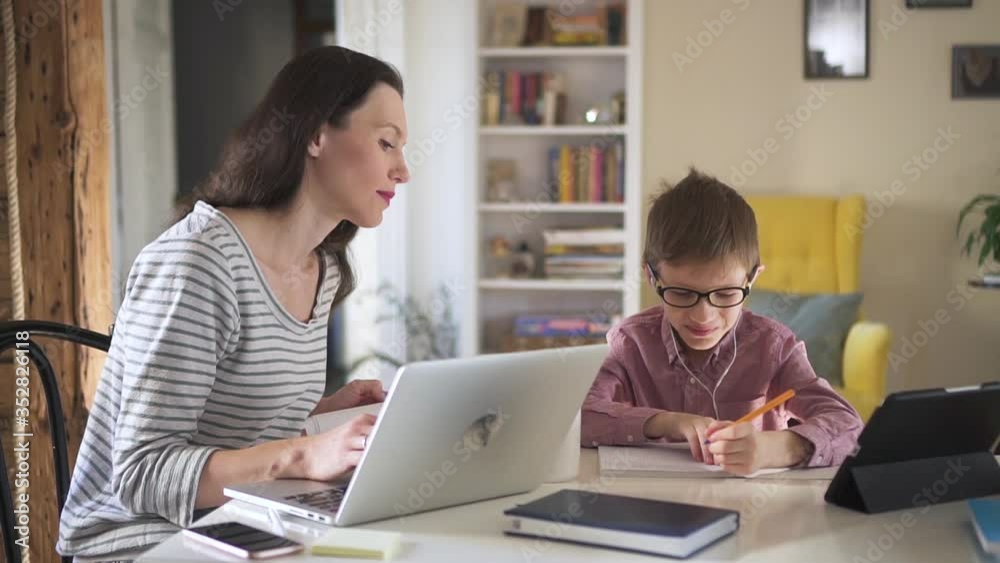 American woman works with laptop remotely and helps schoolboy in learning at table in home spbd. Young mother is working with computer and talking with her son who writing in notebook during online American woman works with laptop remotely and helps schoolboy in learning at table in home spbd. Young mother is working with computer and talking with her son who writing in notebook during online