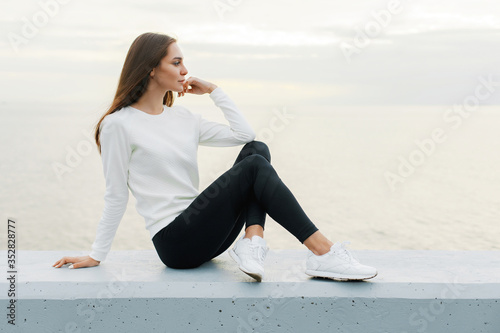 Foto Young woman in white sweater and black leggings is resting after jog on promenad