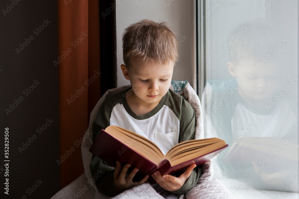Cute little boy reading book while sitting on window sill. Cute little boy reading book while sitting on window sill.