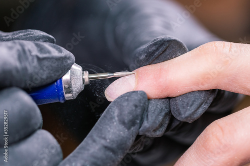 Close-up professional beautician hands working with electric drill on client fingernails. Procedure applying artificial fingernails. Hands of manicurist with tool. Manicurist in gloves removes cuticle