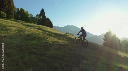 Drone is flying along an athletic man pedalling an MTB E-bike up a steep grassy hill. Beautiful view of the mountains at sunrise/sunset with sun flare. Alone in nature, thinking about life.