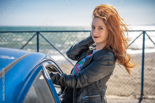beautiful redheaded girl showing half her body through the window of her car with the sea in the background and the wind moving her hair
