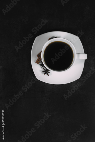 White cup with coffee and saucer, with anise star and cinnamon sticks, on dark textured background. A good idea for the menu or signboard of a restaurant or cafe, cafeteria. Top view, space for text