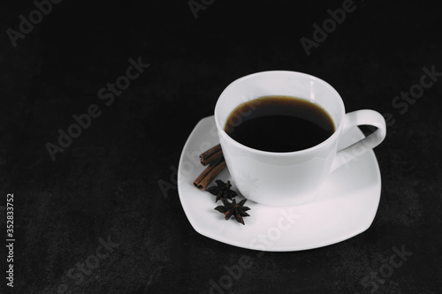 White cup with coffee and saucer, with anise star and cinnamon sticks, on dark textured background. A good idea for the menu or signboard of a restaurant or cafe, cafeteria. Side view, space for text