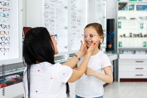 Beautiful little girl in optics store together with mom choose new glasses