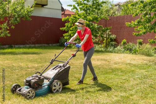 Home life during quarantine or self-isolation. A woman in her backyard mowing grass with a lawn mower on a sunny day at home wearing a surgical mask because of the coronavirus epidemic.