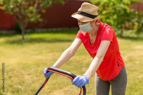 Home life during quarantine or self-isolation. A woman in her backyard mowing grass with a lawn mower on a sunny day at home wearing a surgical mask because of the coronavirus epidemic.