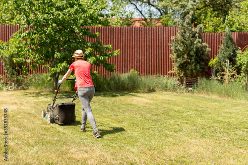 Home life during quarantine or self-isolation. A woman in her backyard mowing grass with a lawn mower on a sunny day at home wearing a surgical mask because of the coronavirus epidemic.