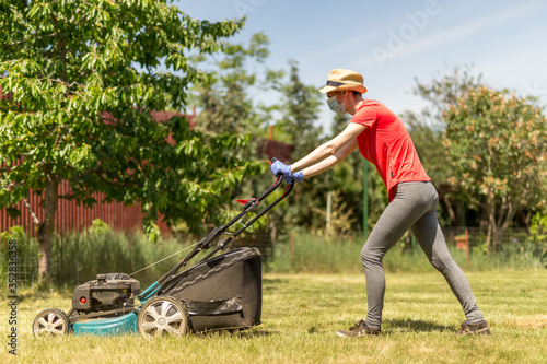 Home life during quarantine or self-isolation. A woman in her backyard mowing grass with a lawn mower on a sunny day at home wearing a surgical mask because of the coronavirus epidemic.