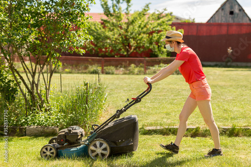 Home life during quarantine or self-isolation. A woman in her backyard mowing grass with a lawn mower on a sunny day at home wearing a surgical mask because of the coronavirus epidemic.