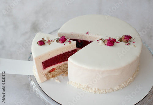 А piece of cake with milk cream mousse and berry filling  is on a white spatula next to the cut cake on a round transparent glass stand. Cake simply decorated with dried rose buds.