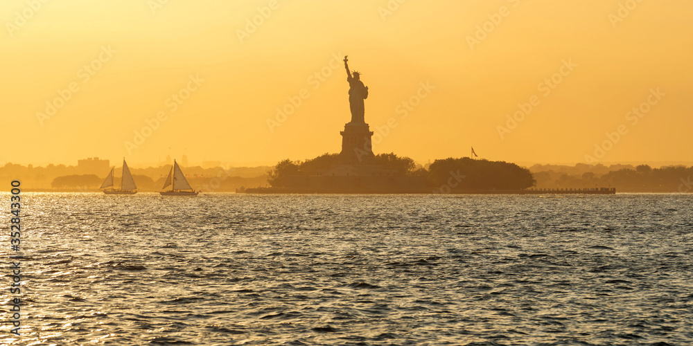 Statue of Liberty and Liberty Island across New York Harbor with sailboats in Summer sunset light. New York City (UNESCO World Heritage)