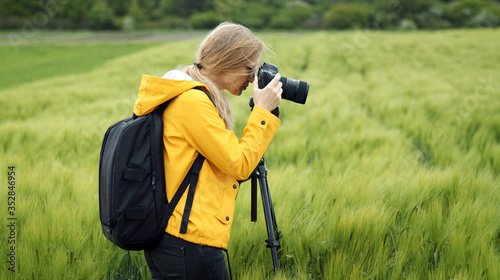 Side view of female photographer standing on spring grain field taking pictures using dslr on tripod