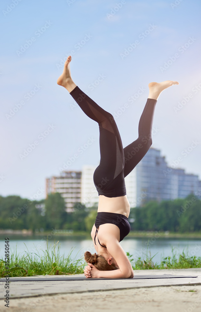 Yoga girl doing asana Shirshasana, standing on her head in the background of urban landscape under a blue sky. Woman working out in black pants and bra. Morning training outdoors at the summer