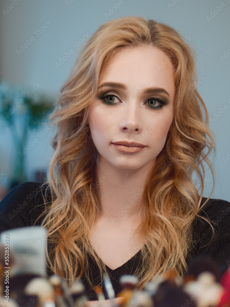 Beautiful blonde sexy young woman with trendy makeup curls hairstyle looking into a mirror reflection at herself liten tungsten light in her domestic rooom Beautiful blonde sexy young woman with trendy makeup curls hairstyle looking into a mirror reflection at herself liten tungsten light in her domestic rooom