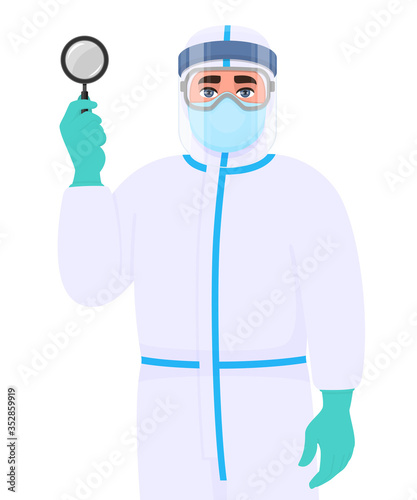 Male doctor in safety protection suit dress, mask, glasses and face shield showing magnifying glass. Physician or surgeon holding magnifier lens. Person wearing personal protective equipment uniform.