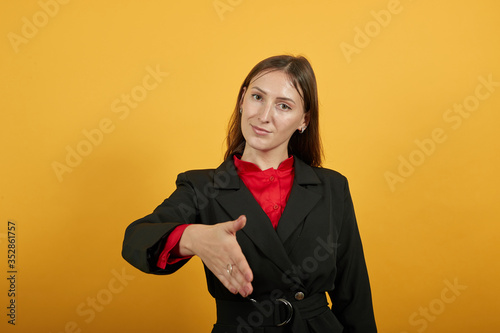 Young Attractive Brunette Woman In Black Stylish Suit, Red Shirt On Yellow Background, Friendly Female Reaches Out To Say Hello. The Concept Of Good People Etiquette, Hospitality