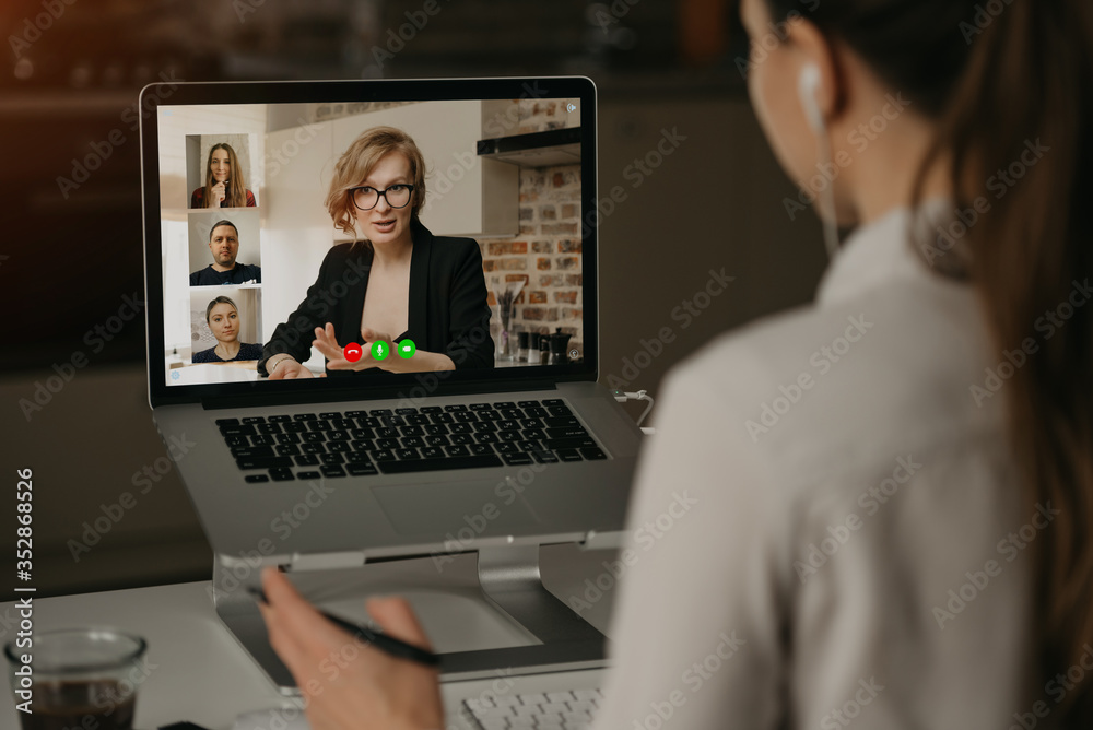 Back view of a woman at home talking with her boss and other colleagues in a video call on a laptop. Businesswoman talks with coworkers on a webcam conference. Business team having an online meeting.