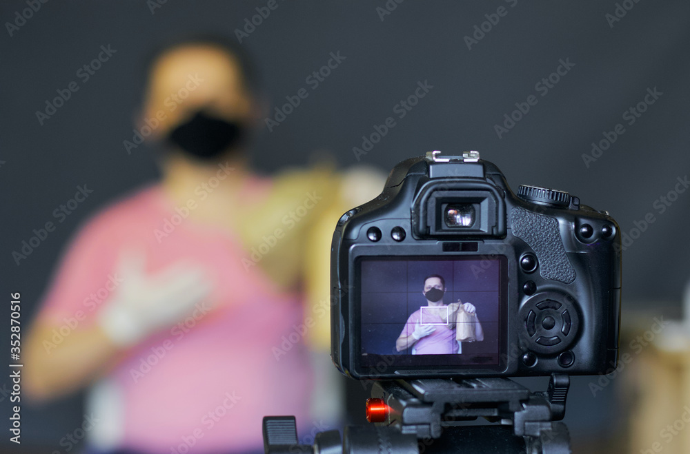 A blogger in a protective mask takes pictures of himself on camera. Advertises a product. Focus is on his camera on a tripod and the image on it. Everything else is blurry.