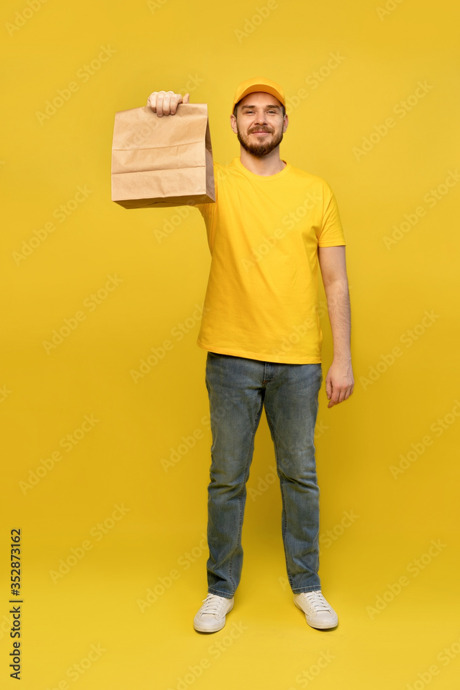 Portrait of young delivery man in yellow uniform with paper packet isolated over yellow background. Portrait of young delivery man in yellow uniform with paper packet isolated over yellow background.